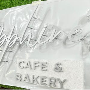 May include: A white sign with the word "Sapphires" in cursive script, and the words "Cafe & Bakery" in block letters. The sign is made of a light-coloured material, likely for a business.
