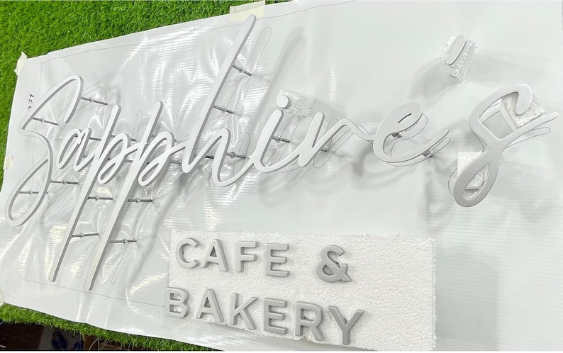 May include: A white sign with the word "Sapphires" in cursive script, and the words "Cafe & Bakery" in block letters. The sign is made of a light-coloured material, likely for a business.