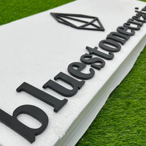 May include: Black sign featuring the text "bluestone creations" and a geometric diamond shape. The sign is on a white surface, with a green background.