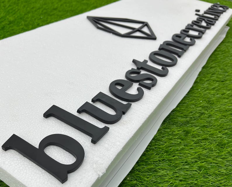 May include: Black sign featuring the text "bluestone creations" and a geometric diamond shape. The sign is on a white surface, with a green background.