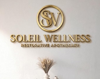 May include: Gold-coloured wall sign for Soleil Wellness, a restorative apothecary. The sign features the initials "SV" within a circle, and the full name is displayed below. Two white vases with a decorative arrangement of dried plants are on a white surface.