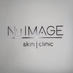May include: The Nu Image Skin Clinic logo is displayed on a white wall. The words "Nu Image" are in a silver, metallic font, with "skin | clinic" in a smaller, black font below. A silver line is positioned under "Nu Image".