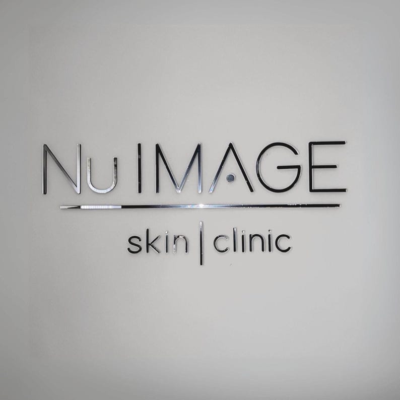May include: The Nu Image Skin Clinic logo is displayed on a white wall. The words "Nu Image" are in a silver, metallic font, with "skin | clinic" in a smaller, black font below. A silver line is positioned under "Nu Image".