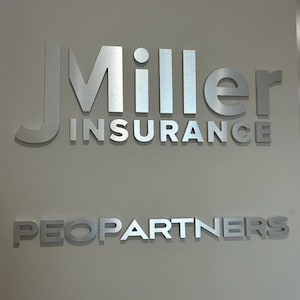 May include: Silver metal signage on a light beige wall. The top line reads "J Miller" with "INSURANCE" below. The bottom line says "PEOPARTNERS" in the same silver metal.