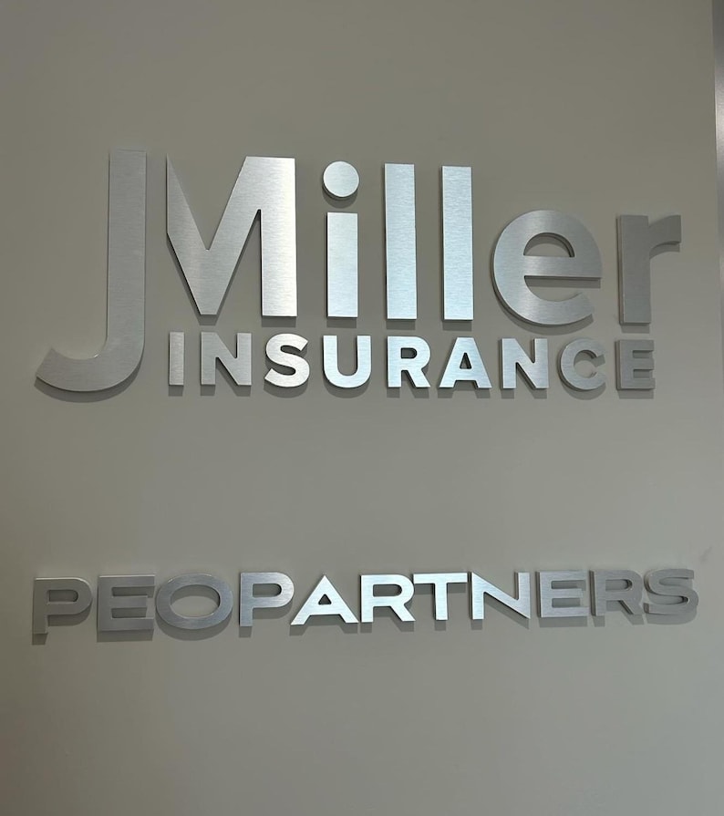 May include: Silver metal signage on a light beige wall. The top line reads "J Miller" with "INSURANCE" below. The bottom line says "PEOPARTNERS" in the same silver metal.
