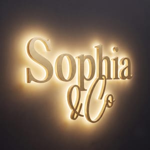 May include: A wall-mounted sign featuring the gold-toned, three-dimensional lettering "Sophia & Co" with a brushed metal finish. The letters are illuminated from behind, creating a soft, warm glow that outlines each character against the wall.