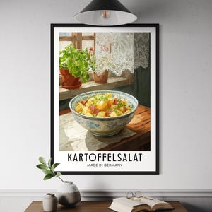 Puede incluir: Una impresión enmarcada que presenta Kartoffelsalat, una ensalada de patatas alemana, en un cuenco con dibujos azules y blancos. La impresión incluye el texto "Kartoffelsalat Made in Germany". La imagen está situada frente a una ventana con plantas y cortinas de encaje.