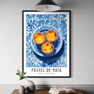 May include: A framed print depicting three Pastel de Nata pastries on a blue and white plate, set against blue and white tiles. The text "PASTEL DE NATA MADE IN PORTUGAL" is below the image. The print is displayed on a wooden table.