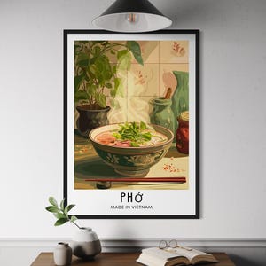 May include: A framed art print featuring a bowl of pho, with steam rising from the broth. The print includes the word "PHO" and "MADE IN VIETNAM" in white text. The artwork has a black frame and is displayed on a white wall.