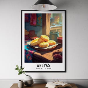 May include: A framed print featuring a still life of arepas, a traditional Colombian dish. The image shows several golden-brown arepas on a wooden board, with the word "AREPAS" and "MADE IN COLOMBIA" printed below. The scene is set on a wooden table.