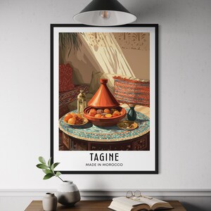 Może przedstawiać: Oprawiony wydruk przedstawiający ilustrację marokańskiego dania tagine wypełnionego jedzeniem, ustawionego na zdobionym stole. Wydruk zawiera słowo "TAGINE" i "MADE IN MOROCCO". Scena skąpana jest w ciepłym świetle, co przywołuje bogactwo kulturowe.