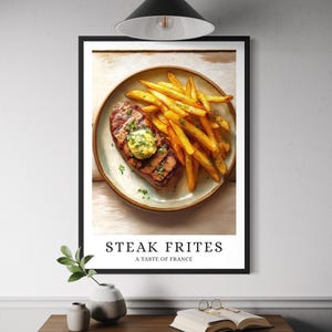 May include: A framed print featuring a steak with a pat of butter and a side of golden french fries on a plate. The text "STEAK FRITES A TASTE OF FRANCE" is displayed below the image. The print is displayed on a white wall.