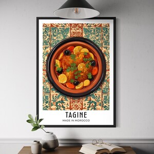 Może przedstawiać: Oprawiony wydruk przedstawiający ilustrację dania tagine. Grafika przedstawia okrągłe, gliniane naczynie wypełnione jedzeniem, udekorowane oliwkami, cytrynami i ziołami. Wydruk zawiera słowo "TAGINE" i "MADE IN MOROCCO" w nowoczesnej czcionce.