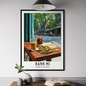 May include: A framed print featuring a vibrant street scene with a table set for a meal. Two sandwiches, two drinks, and the text "BÁNH MÌ MADE IN VIETNAM" are visible. The background shows a street with trees and motorcycles.