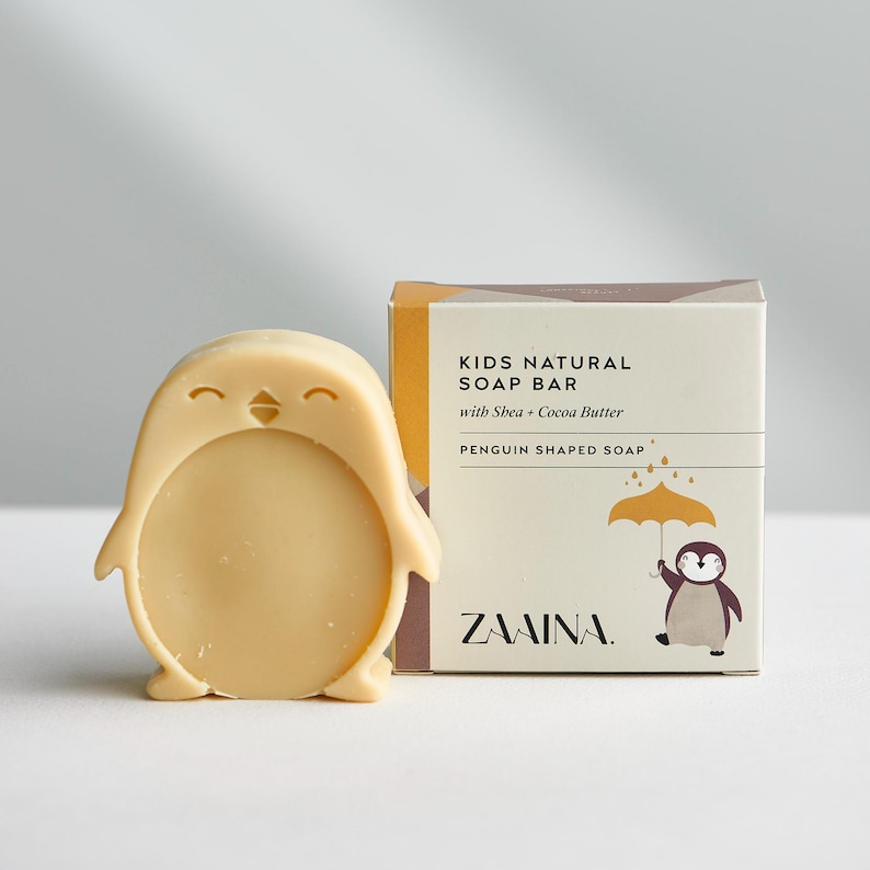 May include: A light beige penguin-shaped soap bar next to its square cardboard packaging. The box features the text KIDS NATURAL SOAP BAR with Shea + Cocoa Butter, PENGUIN SHAPED SOAP, and ZAAINA, along with a graphic of a penguin holding an umbrella.