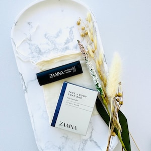 May include: A white marble tray with a black and white soap bar and a black lip balm tube. The soap bar is labeled "FACE + BODY SOAP BAR with Lavender 1.2 OZ / 34 GM ZAINA."