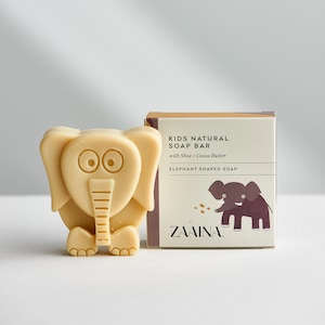 May include: A light beige elephant-shaped soap bar next to its square cardboard packaging. The box features the text KIDS NATURAL SOAP BAR with Shea + Cocoa Butter, ELEPHANT SHAPED SOAP, and ZAAINA, along with a brown elephant illustration.