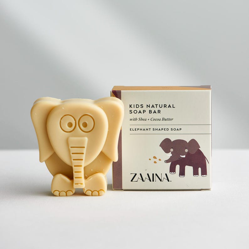 May include: A light beige elephant-shaped soap bar next to its square cardboard packaging. The box features the text KIDS NATURAL SOAP BAR with Shea + Cocoa Butter, ELEPHANT SHAPED SOAP, and ZAAINA, along with a brown elephant illustration.