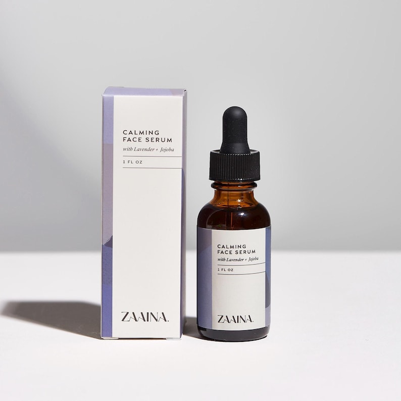 K&ouml;nnte beinhalten: Eine braune Glastropfflasche und die dazugeh&ouml;rige rechteckige Pappschachtel, beide beschriftet mit Calming Face Serum with Lavender + Jojoba, 1 fl oz, von Zaaina. Die Verpackung hat ein wei&szlig;es Etikett mit violetten Akzenten und schwarzem Text.