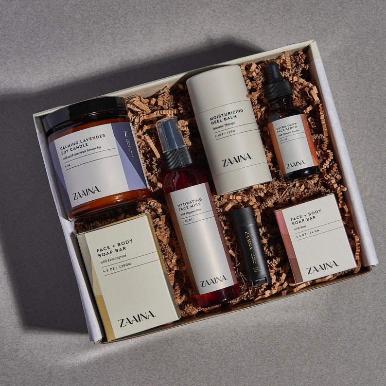 Peut inclure: Une bo&icirc;te cadeau blanche remplie de papier brun et d'une vari&eacute;t&eacute; de produits de soins personnels, dont une bougie, une brume pour le visage, un savon, un baume &agrave; l&egrave;vres et un flacon de s&eacute;rum. Les produits sont tous &eacute;tiquet&eacute;s avec la marque "ZAAINA".