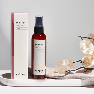 K&ouml;nnte beinhalten: Eine braune Glasflasche mit schwarzer Spr&uuml;hd&uuml;se und einem wei&szlig;en Etikett mit der Aufschrift "Hydrating Face Mist with Organic Aloe 4 FL OZ ZAMINA". Die Flasche steht auf einem wei&szlig;en Marmortablett mit einem beigefarbenen Leinenstoff und einem Zweig mit beigefarbenen Bl&auml;ttern.