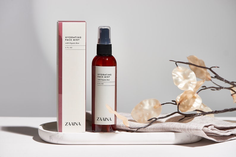 K&ouml;nnte beinhalten: Eine braune Glasflasche mit schwarzer Spr&uuml;hd&uuml;se und einem wei&szlig;en Etikett mit der Aufschrift "Hydrating Face Mist with Organic Aloe 4 FL OZ ZAMINA". Die Flasche steht auf einem wei&szlig;en Marmortablett mit einem beigefarbenen Leinenstoff und einem Zweig mit beigefarbenen Bl&auml;ttern.