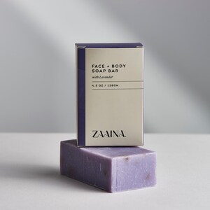 May include: A purple bar of soap with lavender in it, sitting on top of a box that says "FACE + BODY SOAP BAR with Lavender 4.5 OZ / 128GM ZAAMINA."
