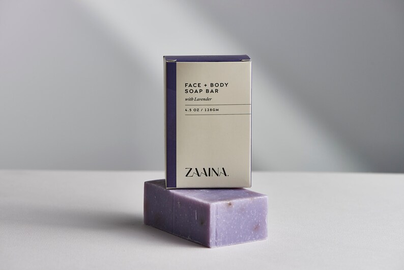 May include: A purple bar of soap with lavender in it, sitting on top of a box that says "FACE + BODY SOAP BAR with Lavender 4.5 OZ / 128GM ZAAMINA."
