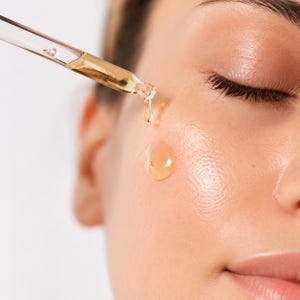 K&ouml;nnte beinhalten: Eine transparente Glastropfpipette gibt ein goldfarbenes, fl&uuml;ssiges Serum auf eine Wange ab. Der viskose, bernsteinfarbene &Ouml;ltropfen ist in der Luft zu sehen, w&auml;hrend er von der Pipettenspitze auf die hydratisierte und gl&auml;nzende Haut f&auml;llt.