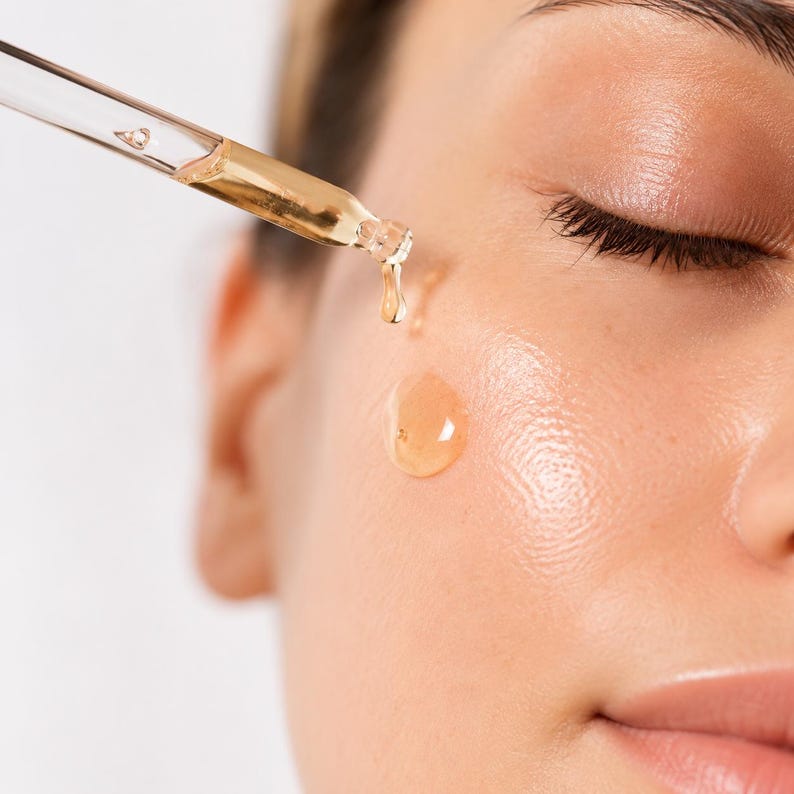 K&ouml;nnte beinhalten: Eine transparente Glastropfpipette gibt ein goldfarbenes, fl&uuml;ssiges Serum auf eine Wange ab. Der viskose, bernsteinfarbene &Ouml;ltropfen ist in der Luft zu sehen, w&auml;hrend er von der Pipettenspitze auf die hydratisierte und gl&auml;nzende Haut f&auml;llt.