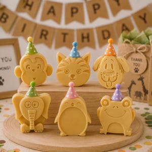 May include: Six light yellow animal-shaped soaps arranged on a wooden block. Each soap features a different animal: a monkey, cat, dog, elephant, penguin, and frog. Every animal wears a small, colorful, polka-dotted party hat on its head.