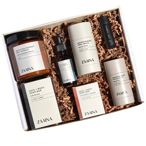 May include: A gift box filled with self-care products, including a soy candle, face serum, hand balm, and two bars of soap. The box is filled with brown paper shreds and the products are all labeled with the brand name "ZAAINA."