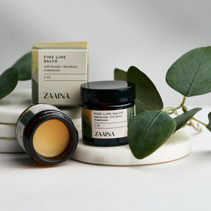 May include: Two jars of fine line salve with avocado, aloe butter, and frankincense. The jars are labeled "ZAAINA" and have a brown label with white text. The smaller jar is 1 oz and the larger jar is 2 oz.