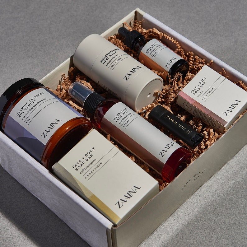 May include: A gift box filled with skincare products, including a candle, face mist, and soap bars. The products are in white and brown packaging with the brand name "ZAAINA". The box is lined with brown paper and sits on a gray surface.