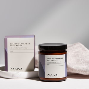 Puede incluir: Una vela de soja en un tarro de vidrio marr&oacute;n con tapa negra y una etiqueta blanca que dice "Calming Lavender Soy Candle with soft American Grown Soy 8 oz ZAINA."
