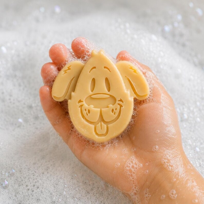 May include: A light tan, dog-shaped bar of soap with a molded facial design featuring floppy ears, a rounded snout, and a small tongue. The soap is covered in white soap bubbles, highlighting the detailed embossed features of the canine face.