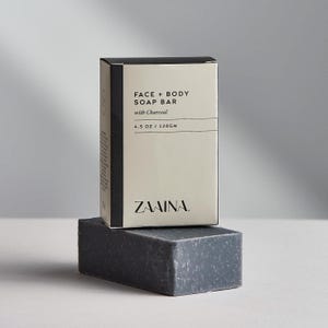 May include: A charcoal face and body soap bar, with a matching box. The box is light beige with black text that reads "FACE + BODY SOAP BAR with Charcoal" and "4.5 OZ / 128GM". The soap bar is dark gray and rectangular. The brand name "ZAAINA" is printed on the box.