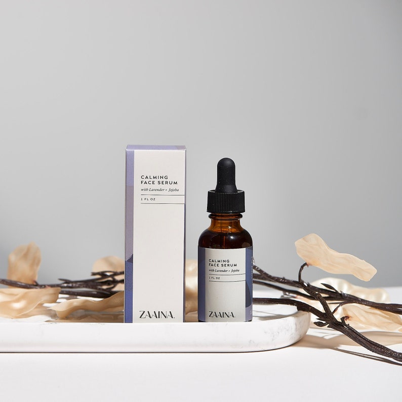 May include: A brown glass bottle of calming face serum with a dropper, labeled "Calming Face Serum with Lavender + Jojoba 1 FL OZ ZAINA". The bottle is sitting on a white marble surface with a branch of dried flowers.