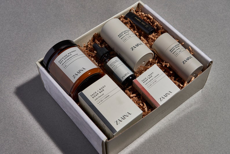 May include: A gift box filled with a variety of self-care products, including a soy candle, face and body soap bar, hand balm, face serum, and lip balm. The box is white with the brand name "Zamina" printed on the side.