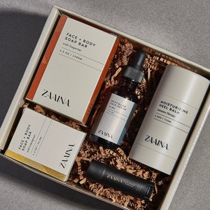 Op de afbeelding: Een cadeaudoos gevuld met bruine papieren strooisel bevat vier ZAAINA huidverzorgingsproducten: een gezichts- en lichaamss zeepreep met mandarijn, een revitaliserend gezichtsserum, een hydraterende hielbalsem en een lippenbalsem. De producten zijn verpakt in witte dozen met zwarte tekst.
