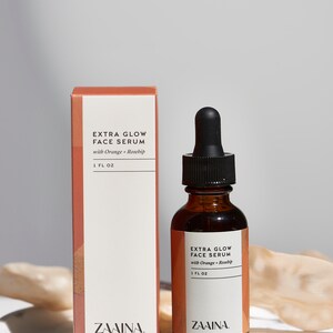 Puede incluir: Un frasco de vidrio de suero facial Extra Glow con naranja y rosa mosqueta, con un tap&oacute;n cuentagotas negro. El frasco est&aacute; sobre una superficie de m&aacute;rmol blanco junto a una caja con la misma etiqueta. El frasco contiene 30 ml de suero.