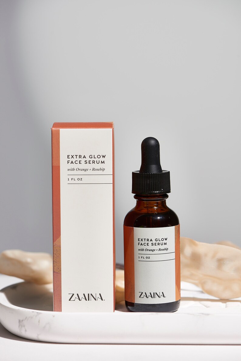 Puede incluir: Un frasco de vidrio de suero facial Extra Glow con naranja y rosa mosqueta, con un tap&oacute;n cuentagotas negro. El frasco est&aacute; sobre una superficie de m&aacute;rmol blanco junto a una caja con la misma etiqueta. El frasco contiene 30 ml de suero.