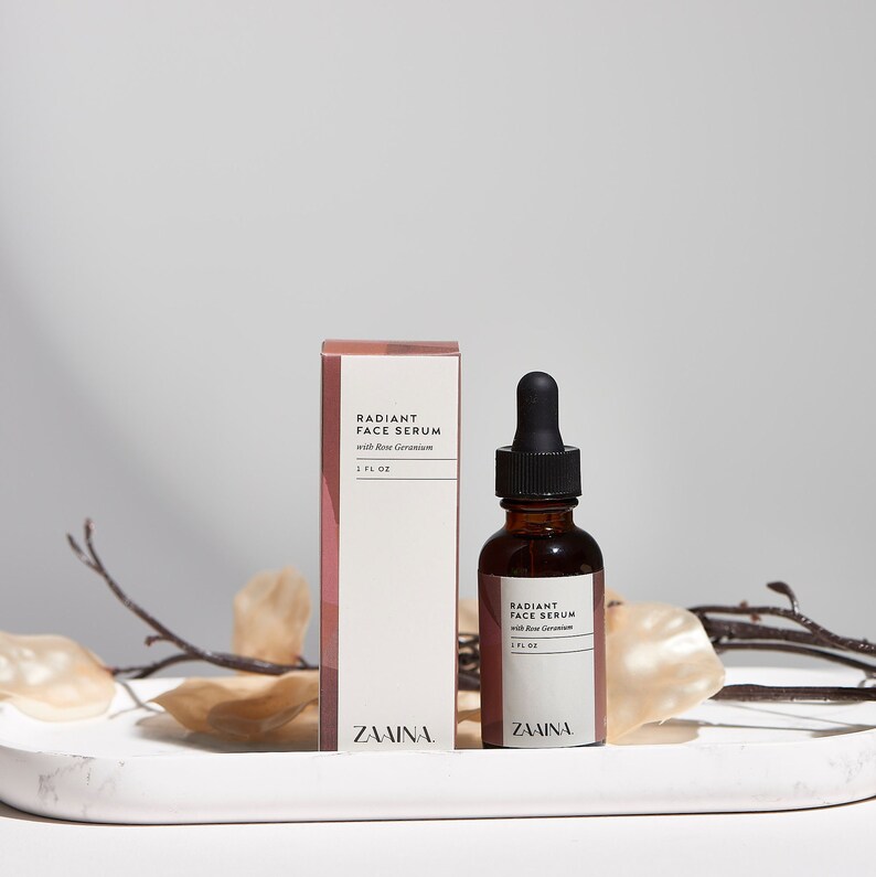 May include: A brown glass bottle with a dropper top filled with a serum labeled "Radiant Face Serum with Rose Geranium 1 fl oz" and the brand name "ZAAINA". The bottle is sitting on a white marble surface next to a box with the same label.