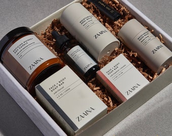 Puede incluir: Una caja de regalo blanca llena de una variedad de productos de cuidado personal, que incluyen una vela de soja, una pastilla de jabón para la cara y el cuerpo, un bálsamo para manos, un bálsamo para talones y un suero facial. La caja está etiquetada con "ZAAINA" y los productos están todos en un empaque blanco y marrón. El texto "BESTSELLER" está impreso en la parte inferior de la caja.