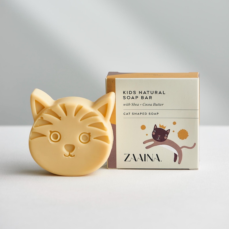 May include: A light beige cat-shaped soap bar next to its square cardboard packaging. The box features the text KIDS NATURAL SOAP BAR with Shea + Cocoa Butter, CAT SHAPED SOAP, and ZAAINA, along with a graphic of a brown cat wearing a crown.