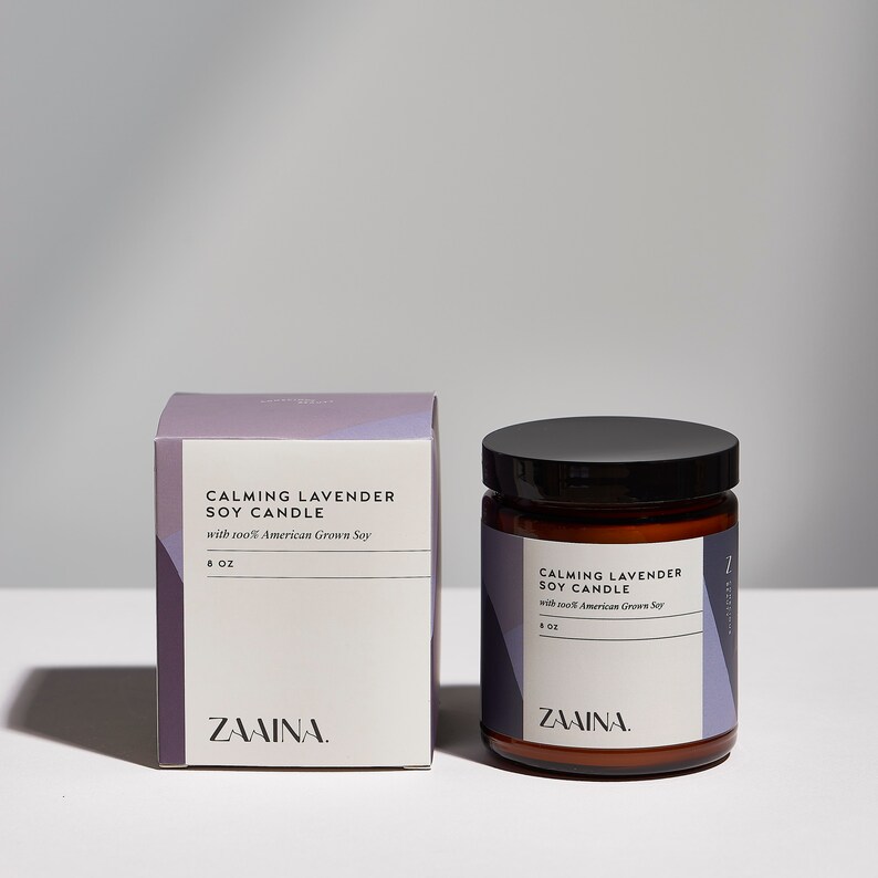 May include: A purple and white box and a glass jar with a black lid. Both containers have the text "Calming Lavender Soy Candle" and "with 100% American Grown Soy" printed on them. The box and jar are labeled "ZAAINA."