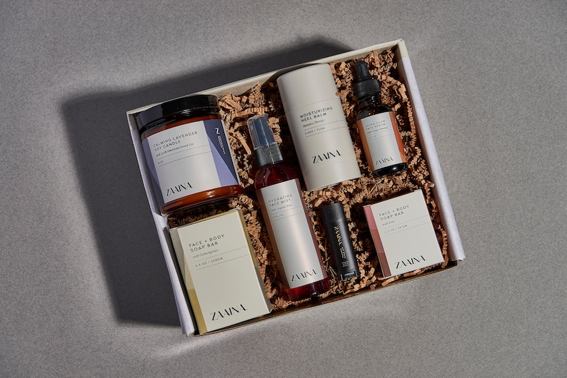 K&ouml;nnte beinhalten: Eine Geschenkbox gef&uuml;llt mit einer Auswahl an K&ouml;rperpflegeprodukten, darunter eine Kerze, ein Gesichtsnebel, eine K&ouml;rpercreme, ein Lippenbalsam und eine Seife. Die Produkte stammen alle von der Marke "ZAINA" und sind in einer wei&szlig;en Box mit braunem F&uuml;llpapier verpackt.