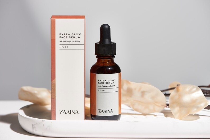 K&ouml;nnte beinhalten: Eine braune Glasflasche mit schwarzem Tropfer und einem wei&szlig;en Etikett mit der Aufschrift "EXTRA GLOW FACE SERUM with Orange + Rosehip 1 FL OZ ZAAINA."