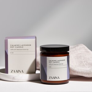 K&ouml;nnte beinhalten: Eine braune Glaskerze in einem Glas mit schwarzem Deckel und einem wei&szlig;en Etikett mit der Aufschrift "Calming Lavender Soy Candle with soft American Grown Soy 8 oz ZAINA."