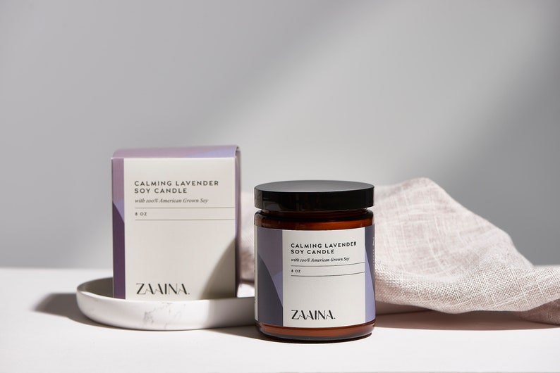 K&ouml;nnte beinhalten: Eine braune Glaskerze in einem Glas mit schwarzem Deckel und einem wei&szlig;en Etikett mit der Aufschrift "Calming Lavender Soy Candle with soft American Grown Soy 8 oz ZAINA."